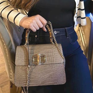 Puede incluir: Un bolso de mano color topo con un estampado de piel de cocodrilo, asas de bambú y un cierre dorado. El bolso tiene una correa de cadena dorada. La persona lleva un top negro y una rebeca color crema con rayas negras.