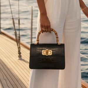 May include: A black handbag with a textured crocodile-embossed finish, featuring a curved bamboo handle and a gold-toned metal turn-lock closure. The bag is rectangular in shape and includes the text STUDIO TEW in the bottom right corner.