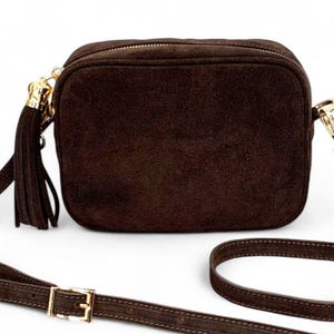 May include: A dark brown suede crossbody bag with a rectangular shape. The bag features a gold zipper, a matching tassel, and a long, adjustable strap with a gold buckle. The bag is designed for everyday use.