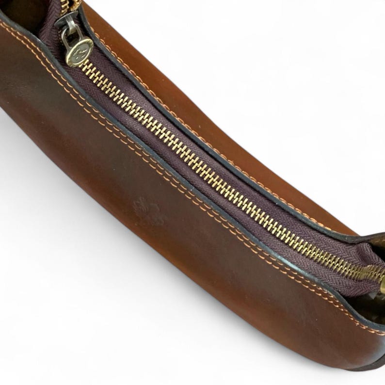 May include: A brown leather pencil case featuring a dark brown zipper closure with a gold-toned metal pull tab. The case has visible stitching along the edges and a small embossed logo on the surface, showcasing a smooth, structured design.