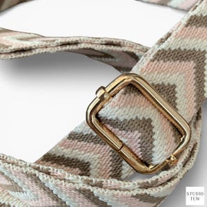 Könnte beinhalten: Nahaufnahme eines gewebten Taschenriemens mit einer goldfarbenen Metallschnalle. Der Riemen weist ein Zickzackmuster in Rosa-, Beige- und Weißtönen auf. Der Riemen ist wahrscheinlich für eine Handtasche.