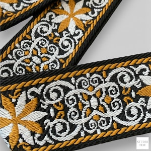 May include: Close-up of a black strap with a detailed pattern. The design features white and gold floral and scrollwork motifs. The edges are trimmed with a gold rope-like border. The strap is likely for a bag or other accessory.