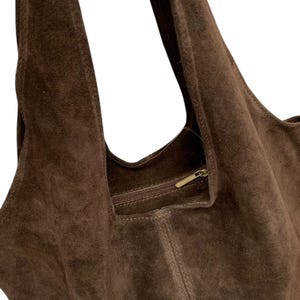 K&ouml;nnte beinhalten: Eine dunkelbraune Hobo-Tasche aus Wildleder mit einer weichen, strukturierten Oberfl&auml;che, einem breiten integrierten Schulterriemen und einer horizontalen Rei&szlig;verschlusstasche im Inneren mit einem metallischen Zipper.