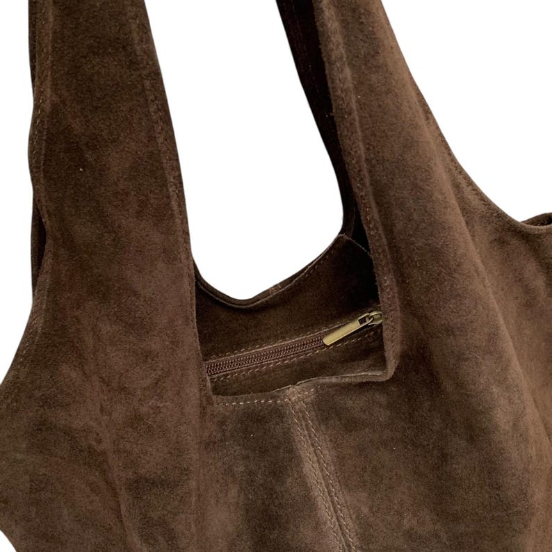 K&ouml;nnte beinhalten: Eine dunkelbraune Hobo-Tasche aus Wildleder mit einer weichen, strukturierten Oberfl&auml;che, einem breiten integrierten Schulterriemen und einer horizontalen Rei&szlig;verschlusstasche im Inneren mit einem metallischen Zipper.