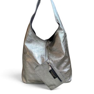 Pode incluir: Uma bolsa hobo prateada metalizada com uma silhueta descontraída e uma alça de ombro única. Presa à bolsa por uma fina corrente prateada, há uma pequena bolsa retangular com zíper e uma etiqueta preta com a inscrição Genuine Leather Made in Italy.