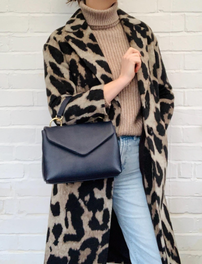 May include: A woman wearing a brown and black leopard print coat, a light brown turtleneck sweater, and light blue jeans. She is carrying a dark blue leather satchel bag with a gold clasp.