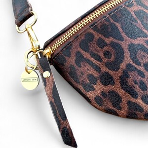 Puede incluir: Un estuche con estampado de leopardo en marr&oacute;n y negro que cuenta con una cremallera de metal dorado, una leng&uuml;eta a juego con el mismo estampado y un dije circular dorado grabado con el texto STUDIO TEW unido a un broche giratorio.