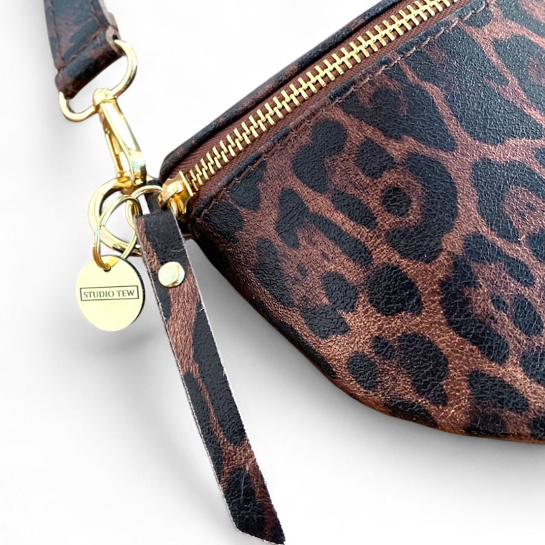 Puede incluir: Un estuche con estampado de leopardo en marr&oacute;n y negro que cuenta con una cremallera de metal dorado, una leng&uuml;eta a juego con el mismo estampado y un dije circular dorado grabado con el texto STUDIO TEW unido a un broche giratorio.