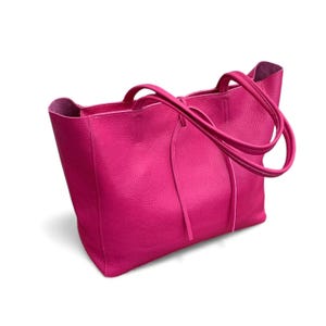 May include: A vibrant hot pink tote bag featuring a textured pebbled surface, an open top design, and two matching shoulder straps. The bag includes thin leather tie closures hanging from the front, providing a simple and functional structure.