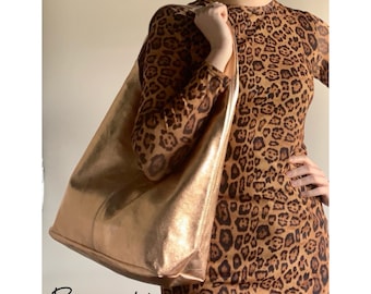 May include: Rose gold leather tote bag with a large, slouchy silhouette. The bag is being held by a person wearing a leopard print dress.