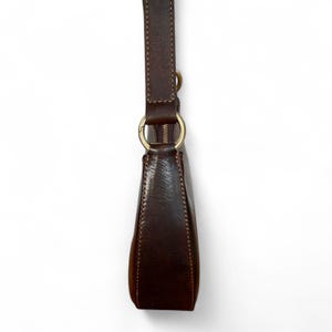 May include: A dark brown leather shoulder bag featuring a smooth texture, visible stitching along the edges, and a gold-toned metal ring connector that attaches the strap to the main body. The bag includes a top zipper closure with gold hardware.