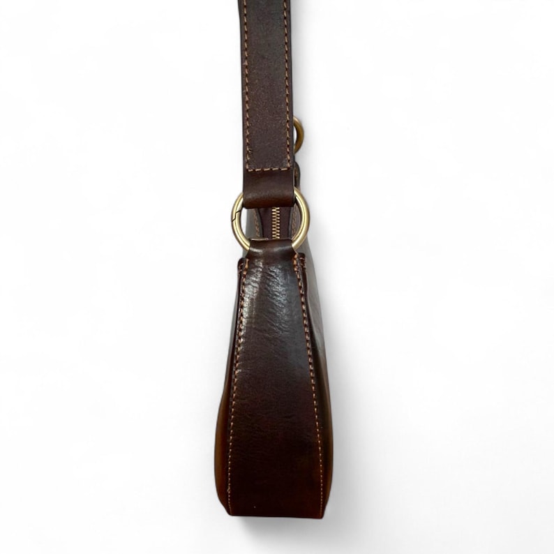 May include: A dark brown leather shoulder bag featuring a smooth texture, visible stitching along the edges, and a gold-toned metal ring connector that attaches the strap to the main body. The bag includes a top zipper closure with gold hardware.