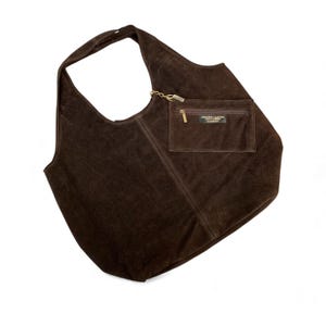 K&ouml;nnte beinhalten: Eine dunkelbraune Wildleder-Hobo-Tasche mit einem einzelnen Schulterriemen und einer befestigten, au&szlig;en liegenden Rei&szlig;verschlusstasche. Die Tasche verf&uuml;gt &uuml;ber einen goldenen Metall-Rei&szlig;verschluss und ein kleines rechteckiges Etikett mit der Aufschrift Genuine Leather Vera Pelle Made in Italy.