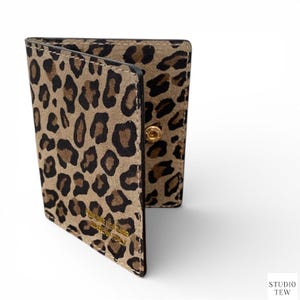 Peut inclure: Un portefeuille pliable avec un motif léopard dans les tons beige et marron. Le portefeuille est ouvert, révélant l'intérieur, et possède une fermeture à bouton-pression dorée. Le texte "STUDIO TEW MADE IN ITALY" est visible sur le devant.