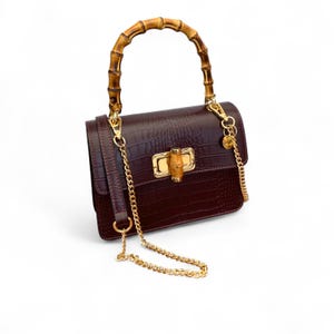 Puede incluir: Un bolso burdeos oscuro con asa de bambú y correa de cadena dorada. El bolso tiene una textura de piel de cocodrilo y un cierre dorado. El asa es de bambú marrón claro.