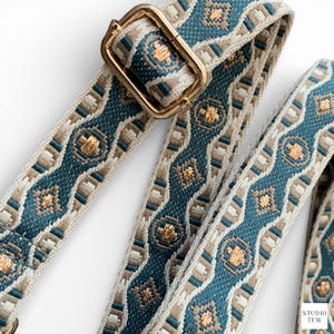 Op de afbeelding: Close-up van een patroon tasriem met een goudkleurige metalen gesp. De riem heeft een herhalend ontwerp in teal, beige en goud. De riem is gemaakt van geweven stof.