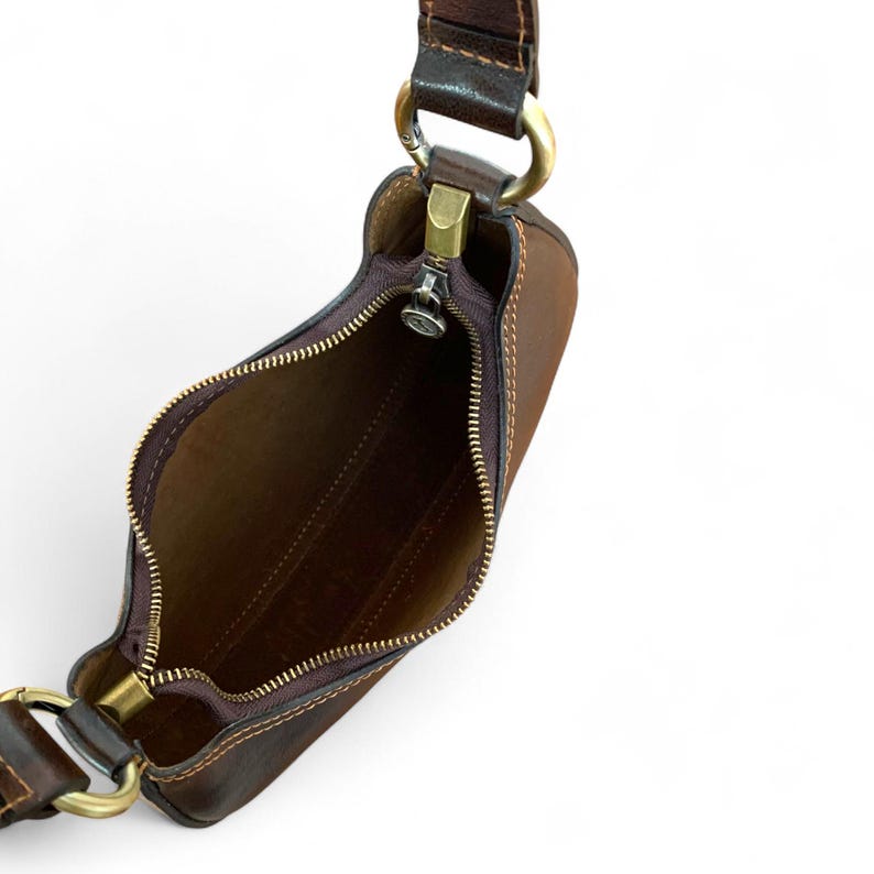 May include: A dark brown leather shoulder bag with a top zipper closure, featuring a smooth interior lining, visible stitching along the edges, and antique brass-toned metal hardware including circular rings that connect the adjustable shoulder strap.
