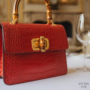 May include: A structured red handbag with a crocodile-embossed texture, featuring a curved bamboo top handle and a gold-toned metal clasp closure with a bamboo accent. The bag has a rectangular flap design and gold hardware attachments for the handle.
