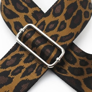 May include: A woven fabric strap featuring a brown and black leopard print pattern. The strap is crossed over itself and secured with a rectangular silver-toned metal slider buckle used for adjusting the length of the strap.