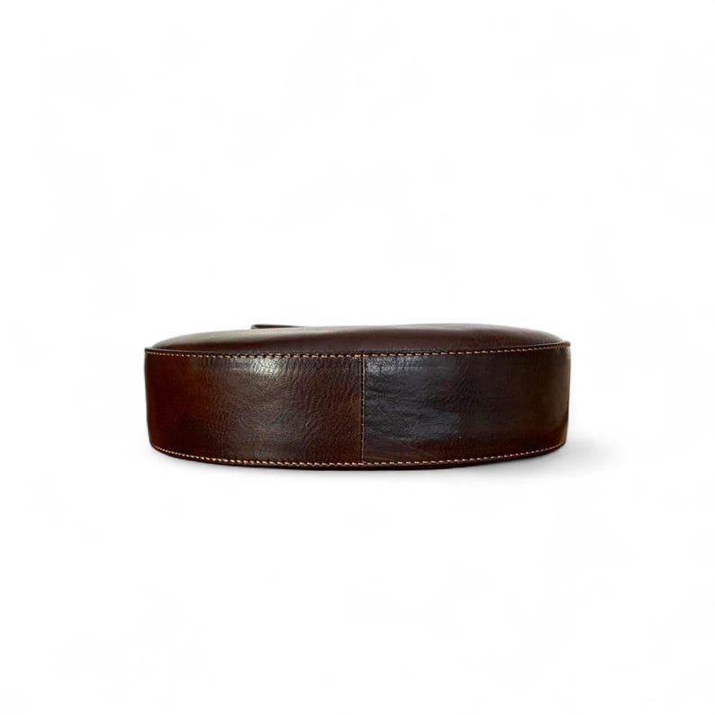 May include: A dark brown leather bag with a smooth, rounded, semi-circular base. The exterior features visible, light-colored stitching along the edges, highlighting the construction of the curved bottom panel against the main body of the bag.