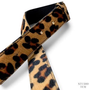 May include: A belt featuring a tan and black leopard print pattern on a textured surface. The belt has a black underside, visible stitching along the edges, and two small gold-toned metal rivets. The text STUDIO TEW is printed in the bottom right corner.