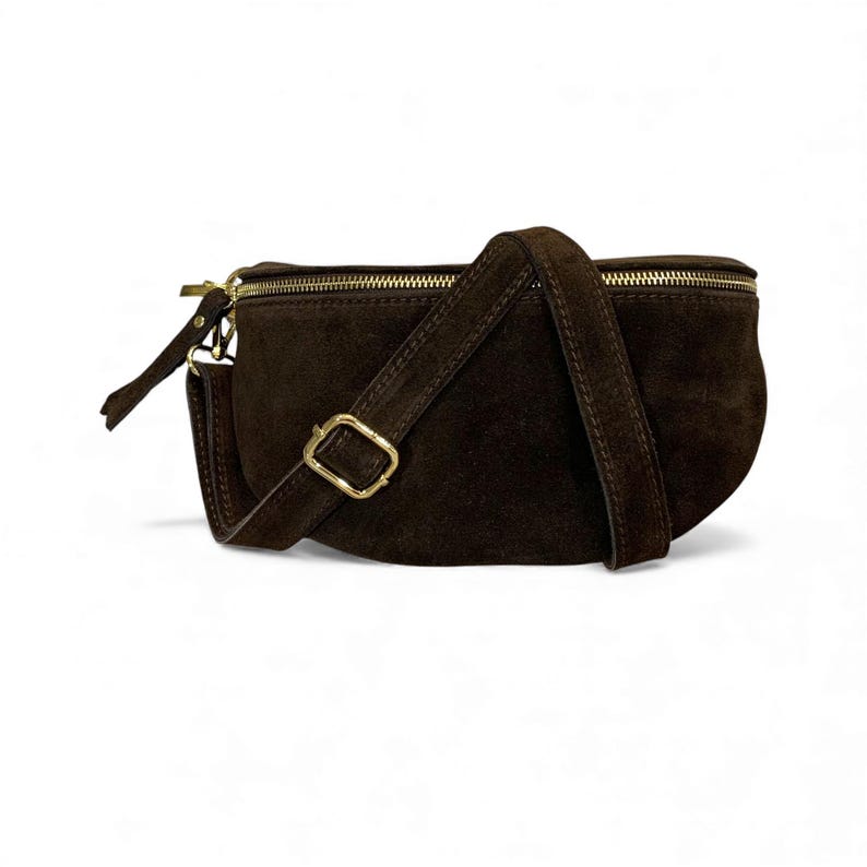 May include: A dark brown suede fanny pack with a gold-colored zipper and buckle. The bag has a curved shape and an adjustable strap. The fanny pack is a stylish accessory for carrying essentials.