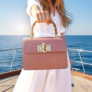 Puede incluir: Un bolso de mano color rosa empolvado con textura de cocodrilo, que cuenta con un cierre de giro met&aacute;lico en tono dorado y un asa de bamb&uacute; curvada. El bolso rectangular estructurado presenta una solapa superior y herrajes pulidos.