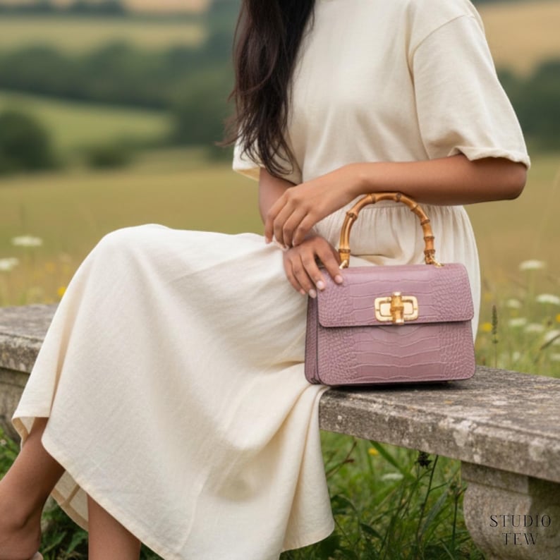 Puede incluir: Un bolso de mano malva con un acabado texturizado en relieve de cocodrilo, que presenta un cierre de giro de metal dorado y un asa superior curva de bamb&uacute;. El bolso rectangular estructurado muestra sus herrajes y material distintivos.