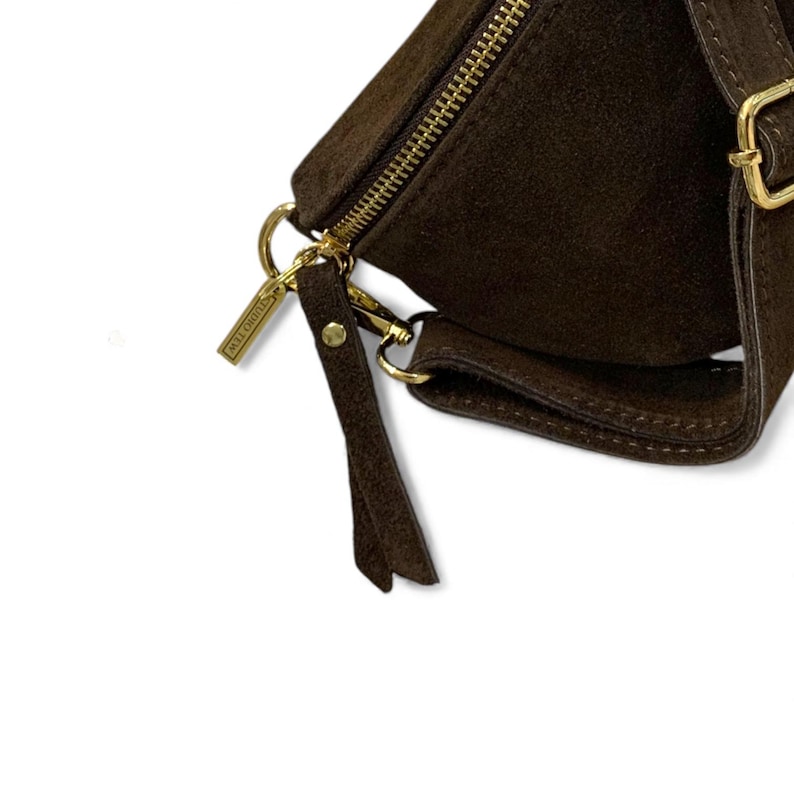 May include: Close-up of a dark brown suede fanny pack with a gold zipper and hardware. The bag has a small, attached pocket and a long, adjustable strap. A small tag with the brand name is attached to the zipper.