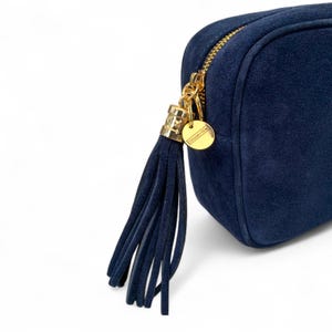 Pode incluir: Uma bolsa transversal de camurça azul marinho com zíper e borla dourados. A bolsa apresenta uma etiqueta dourada Studio 10. A borla é feita de camurça azul marinho. A bolsa tem formato retangular.