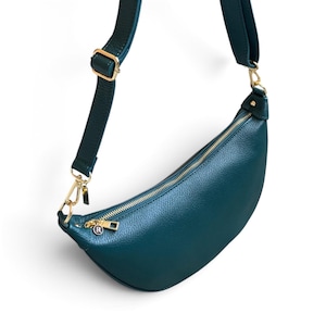 May include: A crescent-shaped teal crossbody bag with a pebbled texture, featuring a gold-tone zip closure, matching gold hardware, and an adjustable shoulder strap. A small circular charm with the letter R is attached to the zip pull.