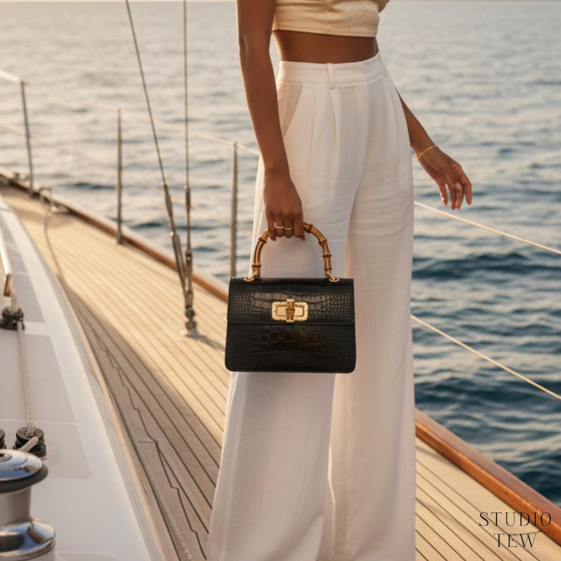 May include: A black handbag with a textured crocodile-embossed finish, featuring a curved bamboo handle and a gold-toned metal turn-lock closure. The bag is rectangular in shape and includes the text STUDIO TEW in the bottom right corner.