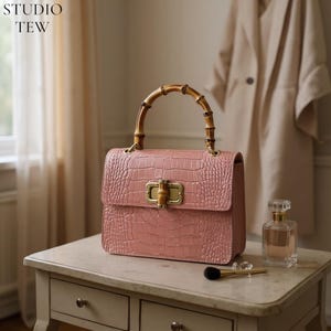 Puede incluir: Un bolso rosa con textura de cocodrilo, asa de bambú y cierre dorado. El bolso está sobre una mesa blanca. El texto "STUDIO TEW" está en la esquina superior izquierda.