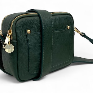 May include: A dark green crossbody bag with a textured leather exterior. The bag features a gold zipper, gold hardware accents, and a matching adjustable strap. A small tag with the text "STUDIO TEW" hangs from the zipper.