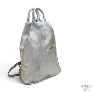 May include: A metallic silver backpack with a structured, tapered silhouette, featuring a top handle, two adjustable shoulder straps, a vertical seam detail down the front, and a small exterior pocket with a subtle embossed logo on the lower right side.