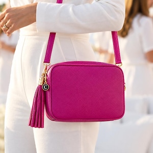 Pode incluir: Uma bolsa tiracolo retangular em rosa fúcsia vibrante com acabamento texturizado semelhante a couro. A bolsa possui uma alça longa combinando, um zíper dourado e um pingente de borla de couro com uma pequena etiqueta preta redonda com um C.
