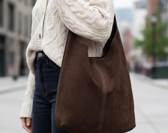 May include: A dark brown suede hobo bag with a slouchy silhouette and a single shoulder strap, worn with a cream-colored cable knit cardigan and dark blue denim jeans. The bag features a vertical seam detail down the center of the front panel.