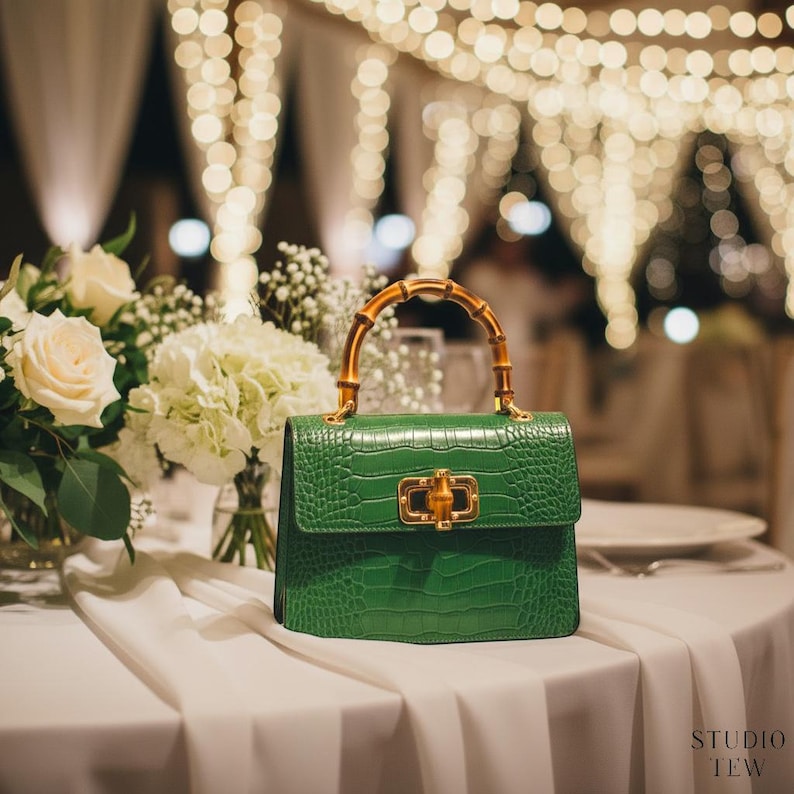 Puede incluir: Un bolso de mano verde estructurado con textura de relieve de cocodrilo, que presenta un cierre de metal dorado y un asa de bamb&uacute; curvada. El bolso tiene una silueta rectangular y est&aacute; colocado sobre una superficie de tela blanca junto a flores blancas.