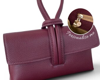 May include: A deep burgundy textured leather clutch bag featuring a single top handle loop and a flap closure. An inset circular detail shows the interior zipper pull with a gold-toned charm engraved with the letter S and the text Personalise me.