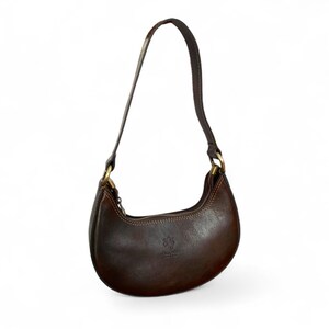 May include: A dark brown leather crescent-shaped shoulder bag with a smooth finish, featuring a single top handle attached with brass-toned metal hardware, a top zipper closure, and visible stitching along the edges of the curved body.