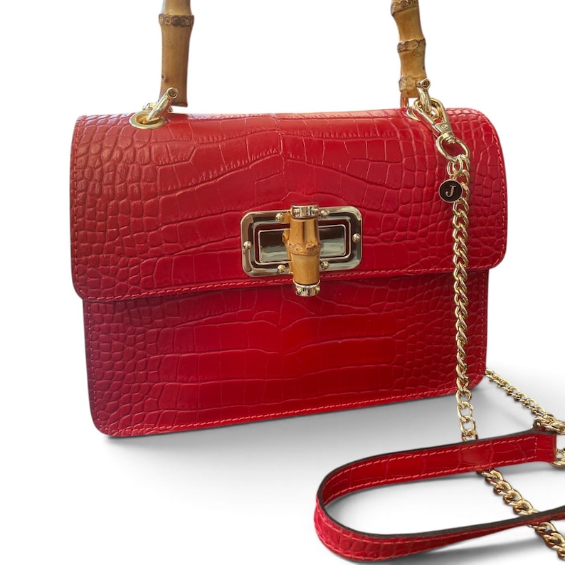 Puede incluir: Un bolso de mano rojo brillante con textura de cocodrilo, que presenta un asa superior de bamb&uacute; y un cierre giratorio de metal dorado estilo bamb&uacute;. El bolso incluye una correa de cadena dorada con un peque&ntilde;o dije circular con la letra J.