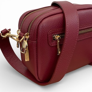 Puede incluir: Bolso bandolera de cuero burdeos con cremalleras y herrajes dorados. El bolso rectangular tiene una correa larga y ajustable. El bolso tiene el texto "STUDIO TEN" en una etiqueta dorada.