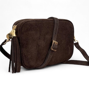 May include: A brown suede crossbody bag with gold-tone hardware. It has a long, adjustable strap and a decorative tassel. The bag is rectangular with a buckle closure on the strap. The bag's dimensions are approximately 20 cm in length.