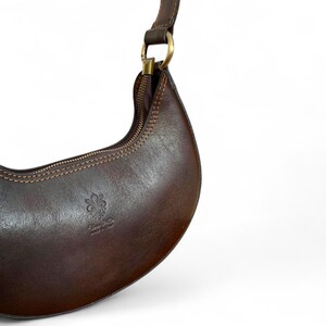 May include: A crescent-shaped dark brown leather shoulder bag featuring a top zipper closure, a gold-toned metal ring attachment for the strap, and a debossed fleur-de-lis emblem with the text Vera Pelle Made in Italy on the front surface.