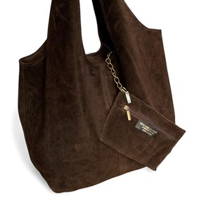 K&ouml;nnte beinhalten: Eine dunkelbraune Wildleder-Hobo-Tasche mit weicher, lockerer Silhouette und einem einzelnen Schulterriemen. Eine kleine passende Wildledertasche ist mit einer goldfarbenen Metallkette befestigt und hat einen Rei&szlig;verschluss sowie ein Etikett.