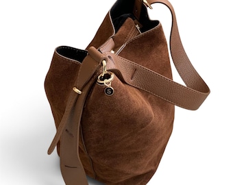 May include: A brown suede bucket bag featuring a smooth leather shoulder strap and a gold-toned metal ring attachment with a small circular charm displaying the letter S. The bag has a structured base and an open top with visible interior lining.