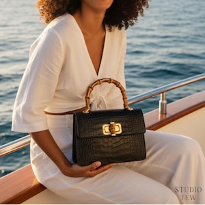 Puede incluir: Un bolso de mano negro con acabado texturizado de piel de cocodrilo, con un asa superior de bamb&uacute; curvado y un cierre de metal dorado con un detalle de bamb&uacute;. El bolso tiene una forma rectangular estructurada y herrajes dorados.