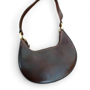 May include: A crescent-shaped dark brown leather shoulder bag featuring a single strap attached with gold-tone metal hardware. The bag has visible light-colored stitching along the edges and a small embossed logo on the front surface.