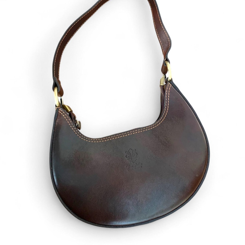 May include: A crescent-shaped dark brown leather shoulder bag featuring a single strap attached with gold-tone metal hardware. The bag has visible light-colored stitching along the edges and a small embossed logo on the front surface.