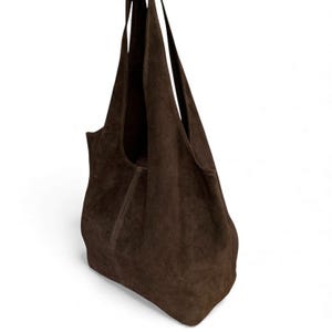 K&ouml;nnte beinhalten: Eine dunkelbraune Hobo-Tasche aus Wildleder mit einer lockeren, unstrukturierten Silhouette. Die Tasche verf&uuml;gt &uuml;ber einen breiten, in den K&ouml;rper integrierten Schulterriemen, sichtbare vertikale Nahtdetails und eine weiche, matte Textur.